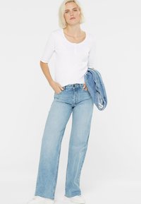 Hellblaue weit geschnittene Jeans mit hoher Taille, kombiniert mit einem taillierten weißen Top mit Dreiviertelärmeln und einem Knopfdetail. Eine Jeansjacke über dem Arm drapiert.