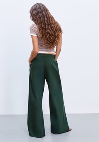 Frau mit langen, welligen Haaren, die ein beiges kurzärmliges Oberteil und eine dunkelgrüne, weite Hose trägt, steht mit den Händen in den Taschen.