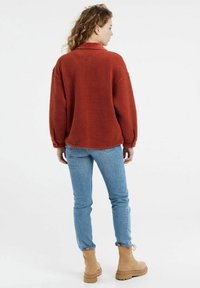 Rust-colored, fuzzy fleece jacket with a collar, long sleeves, and a relaxed fit, paired with light blue jeans and tan lace-up boots.