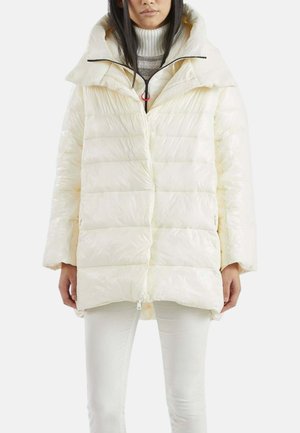 Donna che indossa una giacca imbottita color crema con collo alto sopra un maglione grigio a collo alto e pantaloni bianchi, in piedi davanti a uno sfondo semplice.