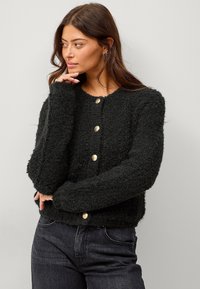 Schwarzer strukturierter Cardigan mit langen Ärmeln, rundem Ausschnitt und fünf goldfarbenen Knöpfen auf der Vorderseite. Kombiniert mit dunklen Jeans.