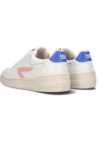 Pár bílých nízkých tenisek s béžovými podrážkami, růžovým bočním detailem a modrými patkami s označením "HUB," zobrazených ze zadního a bočního úhlu.