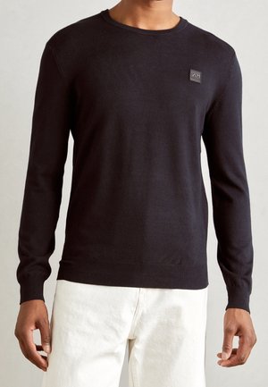 Homme portant un pull noir uni à manches longues avec un petit logo carré sur la poitrine et un pantalon blanc.