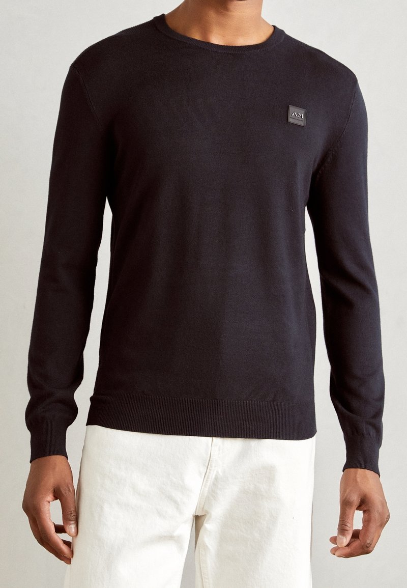 Homme portant un pull noir uni à manches longues avec un petit logo carré sur la poitrine et un pantalon blanc.