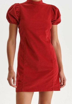 Vrouw die een korte rode fluwelen jurk draagt met opgeblazen korte mouwen en een hoge kraag, staand tegen een effen lichte achtergrond.