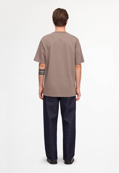 Homme aux cheveux courts et bruns portant un t-shirt taupe oversized, un jean en denim foncé et des chaussures noires, debout de dos avec un bras tatoué visible.