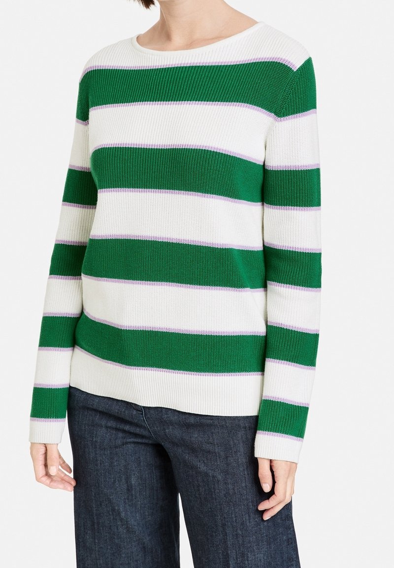 Personne portant un pull en tricot à manches longues avec de larges rayures horizontales vertes et blanches et un jean bleu foncé.