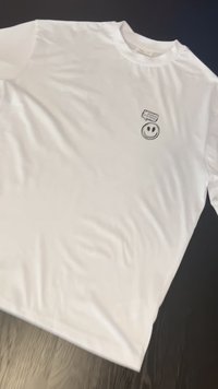Camiseta de algodón blanca con mangas cortas, con un pequeño gráfico de una cara sonriente negra en el lado izquierdo del pecho. Textura suave, escote redondo estándar.