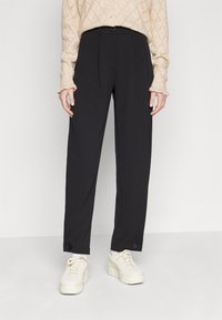 Black trousers with a high waist and front pleats, featuring button accents at the hem, paired with chunky white sneakers.