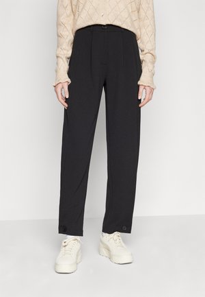 Black trousers with a high waist and front pleats, featuring button accents at the hem, paired with chunky white sneakers.
