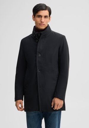 Mann, der einen schwarzen, geknöpften Mantel über einem dunklen Rollkragenpullover und blauen Jeans trägt, steht vor einem einfarbigen hellgrauen Hintergrund.