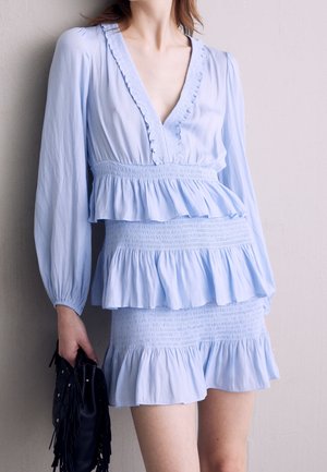 Femme portant une robe bleu clair à manches longues avec des volants et une taille smockée, tenant un sac à main noir à franges contre un mur uni.