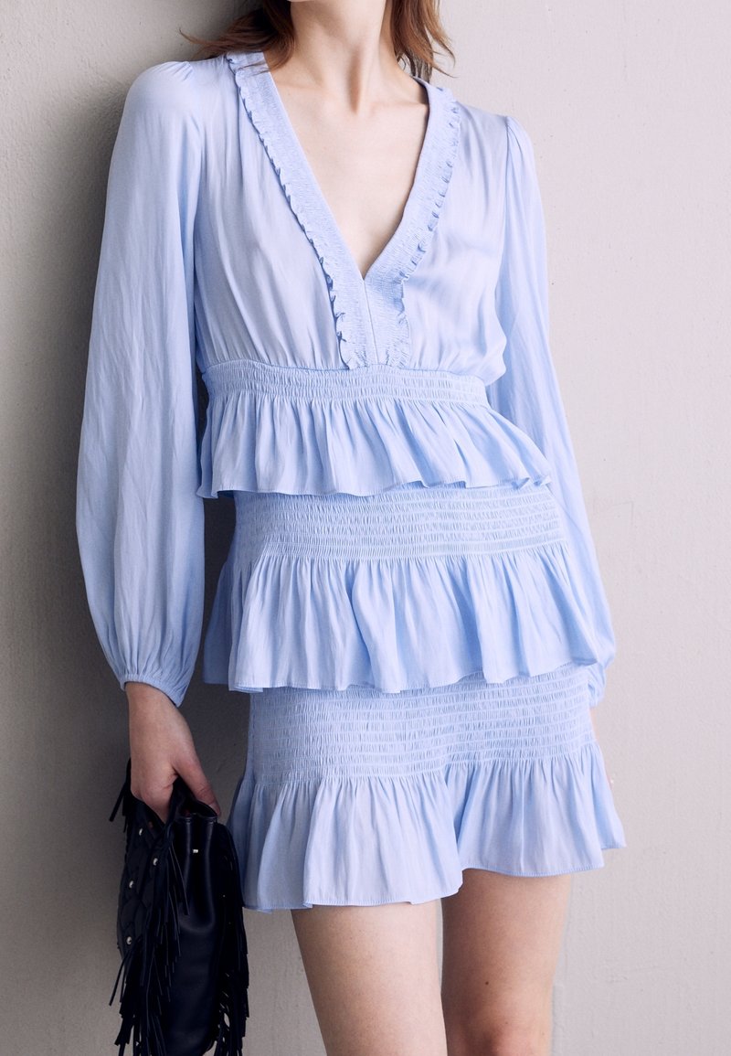 Femme portant une robe bleu clair à manches longues avec des volants et une taille smockée, tenant un sac à main noir à franges contre un mur uni.