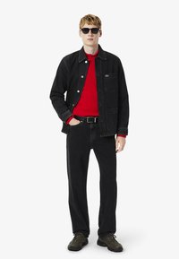 Chaqueta de mezclilla negra con tres bolsillos frontales, suéter de punto rojo abajo, pantalones negros y gafas de sol oscuras. Tejido texturizado con un ajuste casual.