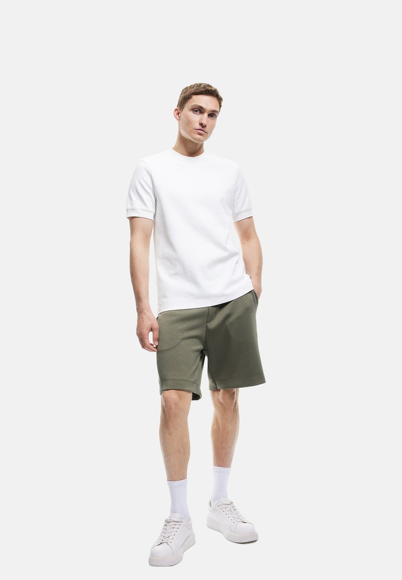 Biała koszulka z krótkim rękawem zestawiona z oliwkowymi szortami, o luźnym kroju. Noszona z białymi skarpetkami i sneakersami. Prosty, casualowy design.