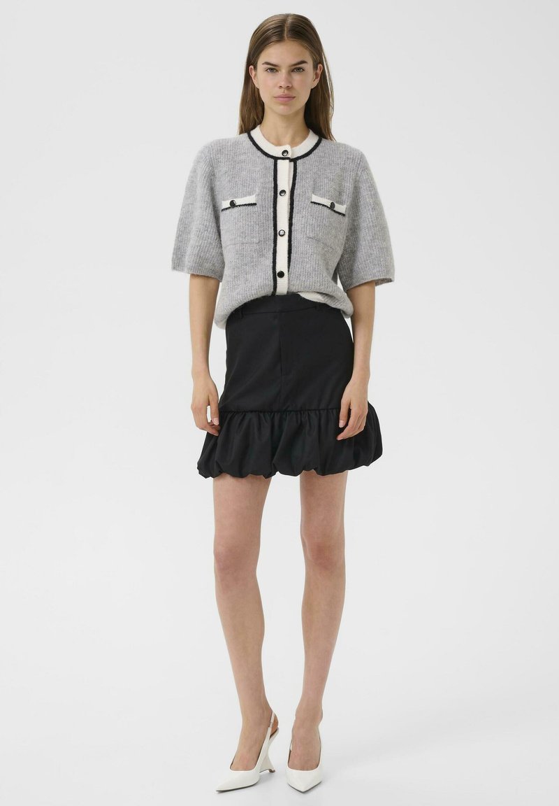 Cardigan gris en tricot avec une bande blanche et des finitions noires, associé à une jupe noire à volants. Le modèle porte des talons blancs pointus.