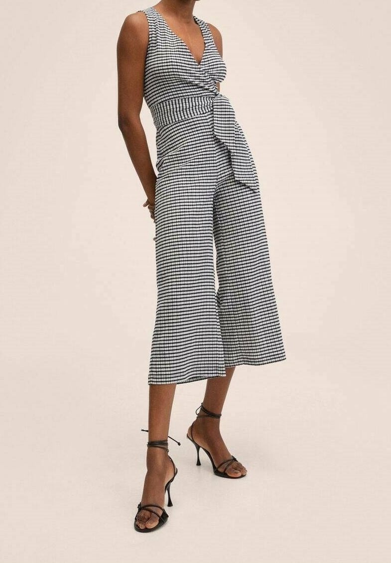 Femme portant une combinaison sans manches à carreaux noir et blanc, avec jambes larges et lien à l'avant, associée à des sandales noires à talons hauts et à lanières.
