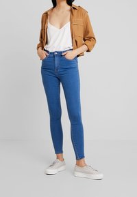 Jeans ajustados azules de cintura alta, tela de denim suave y textura lisa. Combinados con una blusa blanca y una camisa marrón, y zapatillas de color claro.