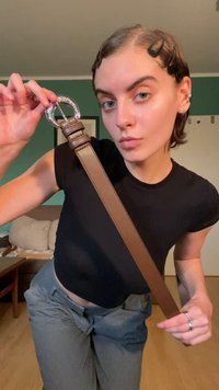 Bruine leren riem met een zilveren ovale gesp, voorzien van gestikte details en een gladde textuur, vastgehouden in een hand tegen een neutrale achtergrond.