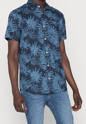 Homme portant une chemise bleu marine à manches courtes avec un motif de feuilles tropicales bleu clair et un jean bleu.
