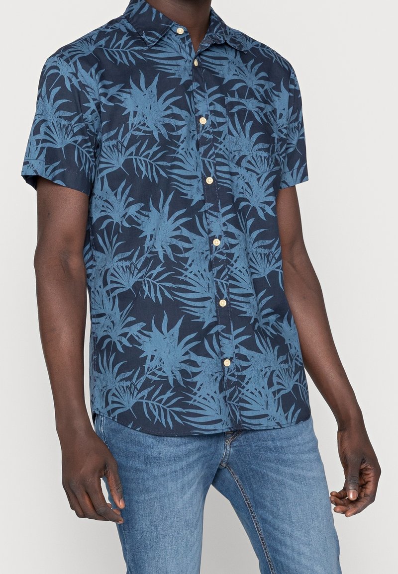 Homme portant une chemise bleu marine à manches courtes avec un motif de feuilles tropicales bleu clair et un jean bleu.