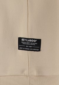 Etiqueta de tela beige que presenta "KEY LARGO" en negro, con texto que detalla la información de la marca y la fecha de establecimiento, cosida sobre un fondo texturizado.