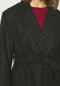 Cappotto in misto lana grigio scuro con design avvolgente, colletto a risvolto e cintura annodata in vita. Camicia rossa a girocollo visibile sotto.