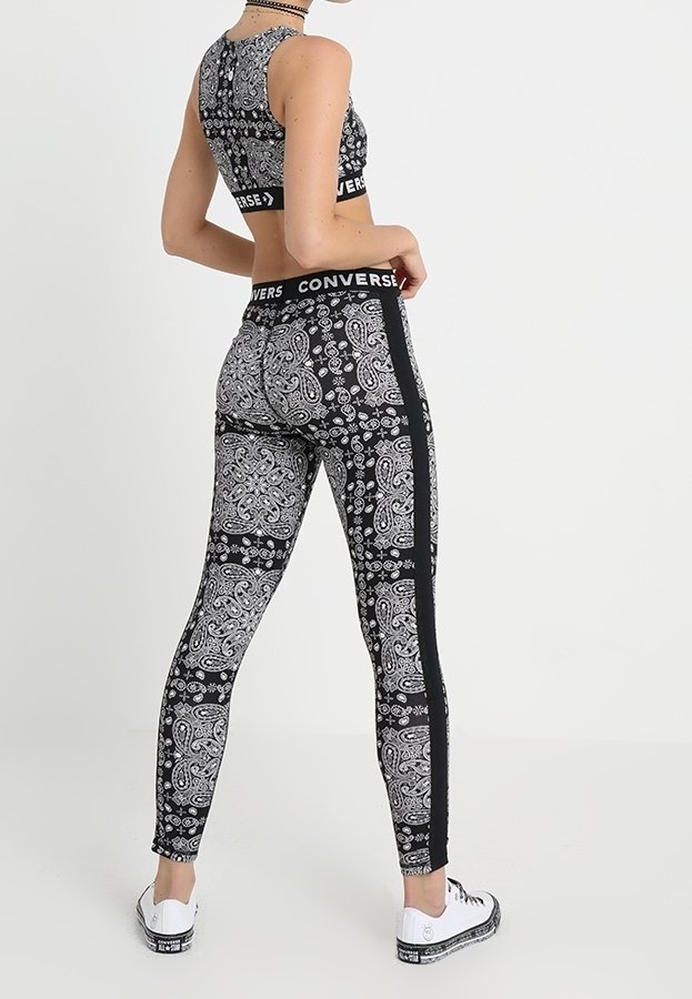 Femme portant des leggings à motif paisley noir et blanc et un crop top assorti avec le logo Converse, debout sur un fond blanc.