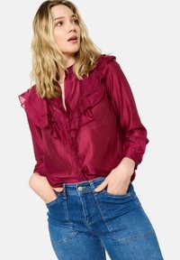 Burgundy button-up blouse with ruffled neckline, paired with high-waisted blue denim jeans. The blouse features a smooth, slightly shiny texture.