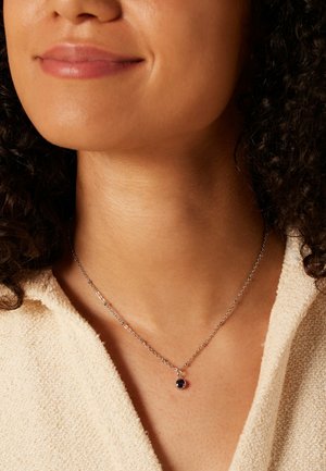 Collier en chaîne argentée avec un pendentif en pierre précieuse ronde et violette, posé sur un vêtement pâle à texture.