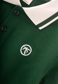 Zelené polo tričko s bielym golierom a pruhom, s logom palma v bielom výšivke. Dvoje čiernych gombíkov na golieri. Textúrovaná tkanina.