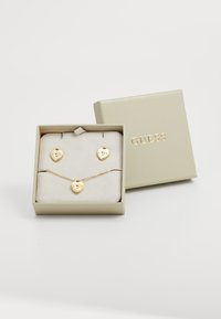 Gold-tone jewelry set in a box, featuring two heart-shaped earrings and a matching heart pendant necklace on a soft beige display cushion.