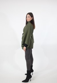 Olijfgroene wikkeljas met brede mouwen, ceintuur in de taille. Gecombineerd met nauwsluitende grijze jeans en zwarte enkellaarsjes. Minimalistisch ontwerp.