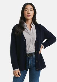 Marinefarbener Cardigan mit gerippter Textur, Knopfverschluss und offener Frontgestaltung, darüber getragen über einem gestreiften Hemd mit Knöpfen und Jeans.