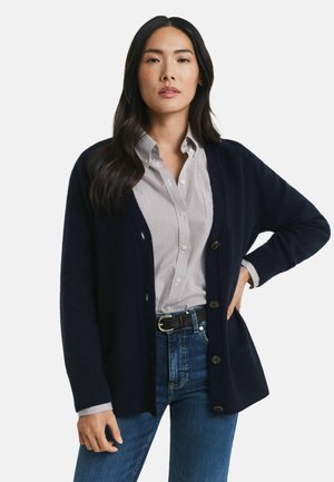Navy cardigan with ribbed texture, button closure, and open front design, layered over a striped button-up shirt and denim jeans.