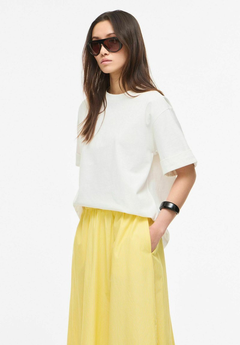 Femme aux longs cheveux bruns, portant des lunettes de soleil foncées, une chemise blanche à manches courtes, une jupe jaune et un bracelet noir, debout avec une main dans la poche.