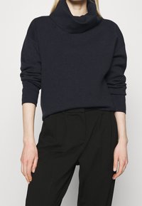 Merihenkinen kauluskaitale-neule pehmeästä kankaasta, pitkähihaiset, yhdistettynä muotonsa mukaan räätälöityihin mustiin housuihin, korostaen rentoa istuvuutta ja modernia muotoilua.