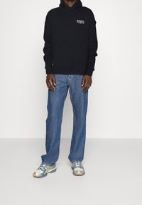 Felpa nera in cotone con stampa "PEQUS", tasca frontale, abbinata a jeans in denim blu e sneakers sportive multicolori.
