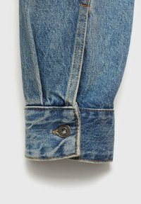 Manga de camisa de mezclilla en azul claro con costuras naranjas en contraste. Presenta un gran botón metálico redondo en la muñeca con una textura suave.