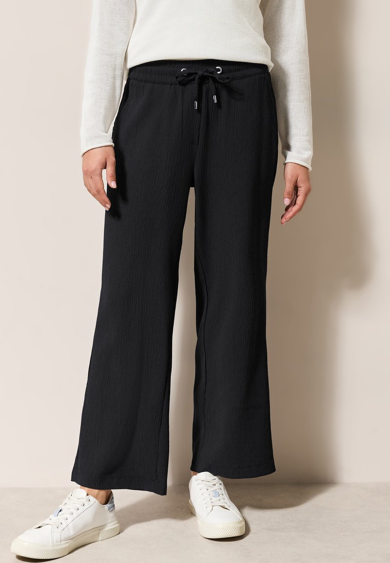 Pantalones de pierna ancha negros con una tela texturizada, cinturilla elástica y cordón ajustable. Llevados con zapatillas blancas.