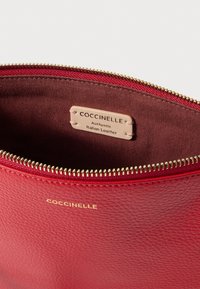 Piros bőrből készült táska texturált felülettel, arany cipzárral és belső márkajelzéssel, amelyen ez áll: "Coccinelle, valódi olasz bőr."