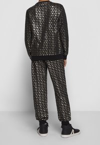 Tuta nera con motivo a logo EA7 oro ripetuto. Design aderente con polsini a coste e supporto alla caviglia. Abbinata a scarpe da ginnastica nere.