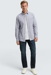 Chemise à carreaux bleu clair à manches longues avec col boutonné, une poche poitrine, portée sur un t-shirt blanc, associée à un jean foncé et des baskets blanches.