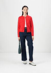 Red cardigan with floral pattern, white top, dark blue wide-leg jeans, and white slip-on shoes. Holding a green handbag with strap.