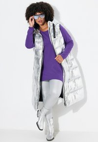 Silberne Tauchweste mit Kapuze, lila langärmliges Oberteil und metallisch silberne Leggings. Silberne Knöchelstiefel runden das Outfit ab.