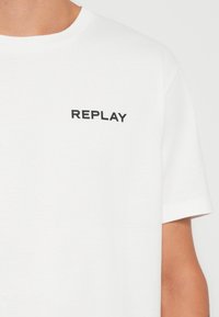 Hvid bomulds t-shirt med korte ærmer, med et sort "REPLAY" logo på venstre bryst. Glat tekstur og klassisk rund hals design.