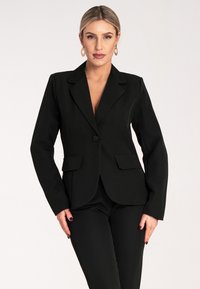 Czarny fasonowany żakiet z pojedynczym zapięciem na guzik, z klapami oraz frontowymi kieszeniami, zestawiony z pasującymi czarnymi spodniami. Gładka tekstura materiału.