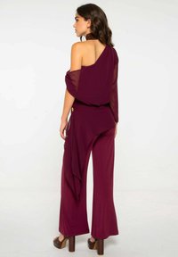 Monosuit burdeos con drapeado en un hombro, tejido suave, piernas anchas y un diseño fluido. Presenta un acento dorado en la cintura. Tacones plataforma marrones.