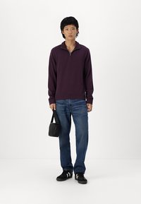 Maglione zip-up viola, in misto cotone, con maniche lunghe, polsini e orlo a costine. Indossato con jeans blu e sneakers nere. Tiene una piccola borsa nera.