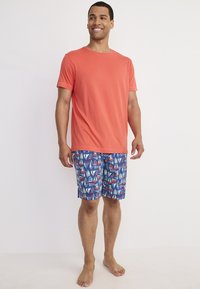 Korallfärgad t-shirt med rund halsringning, kombinerad med blå shorts med färgglada segelbåtsmönster. Modellen står barfota mot en enfärgad bakgrund.